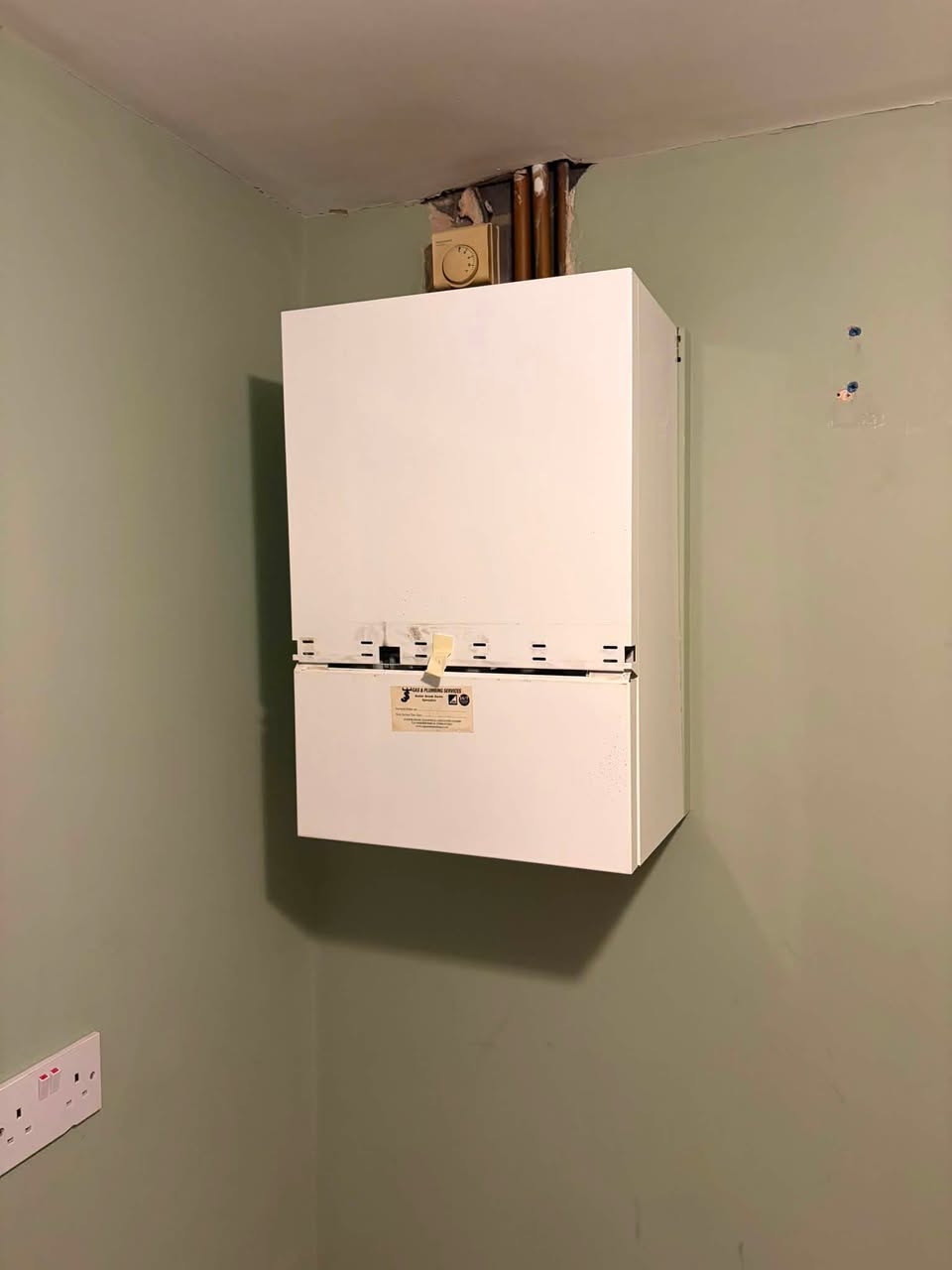 A wall-mounted boiler in the corner of a room, casing on, with copper pipes and a programmer above it.