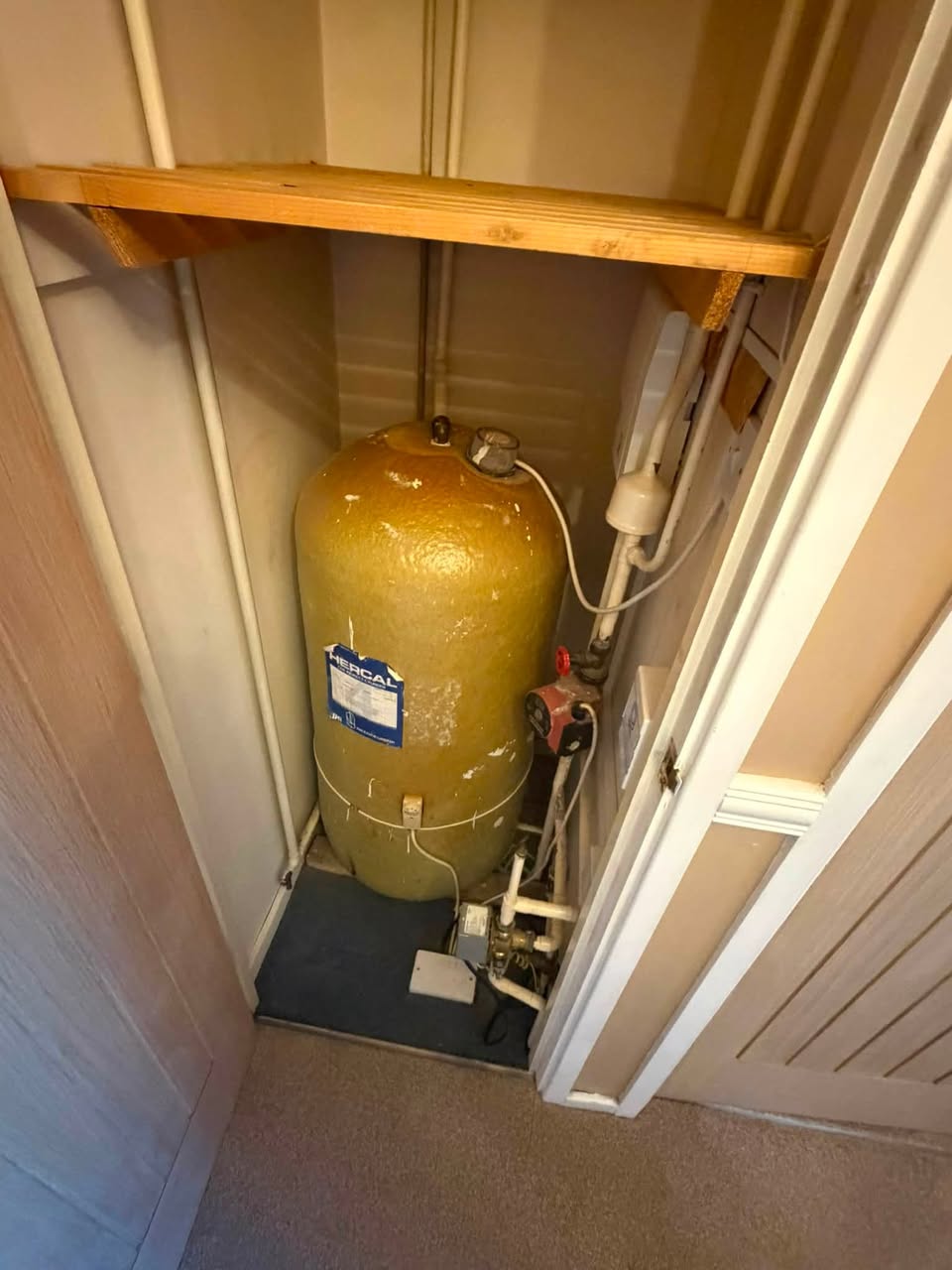 An old foam-lagged hot water cylinder in an airing cupboard with a pump and valves beside it.