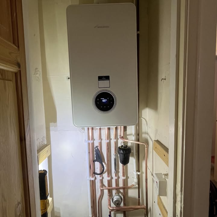 A boiler on a wall with a rank of copper pipes beneath it and the display reading 65 degrees.